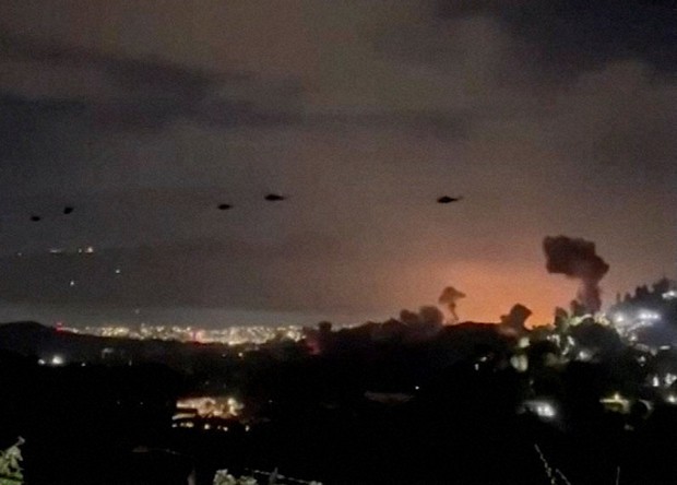 ベネズエラの首都カラカスで爆発により立ち上る煙を背に飛行するヘリ＝2026年1月3日、ロイター