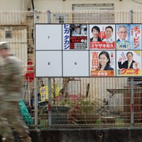 米軍嘉手納基地に隣接する北谷町砂辺地区で、衆院選沖縄2区の候補者ポスター掲示板の前を歩く迷彩服姿の米軍関係者＝沖縄県北谷町で2026年1月30日午前11時36分、喜屋武真之介撮影