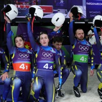 「追悼ヘルメット」の使用で失格となったスケルトンのウクライナ代表、ヘラスケビッチ選手に対し、片膝をついてヘルメットを掲げ敬意を示すウクライナのリュージュ・リレーチーム＝イタリア北部コルティナダンペッツォで2026年2月12日、ロイター