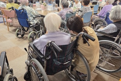 特別養護老人ホームで介護職員（右）に寄り添われる車椅子の高齢者。河口さんは地域全体で支える仕組みづくりの必要性を訴える