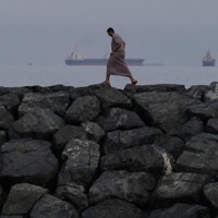 アラブ首長国連邦で、ホルムズ海峡で列をなす原油タンカーや貨物船（奥）と岸辺を歩く男性＝2026年3月11日、AP