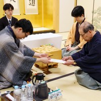 立会の桐山清澄九段（手前右）に封じ手を渡す挑戦者の永瀬拓矢九段（手前左）。奥右は藤井聡太王将＝大阪府高槻市の関西将棋会館で2026年3月25日午後6時8分、西村剛撮影