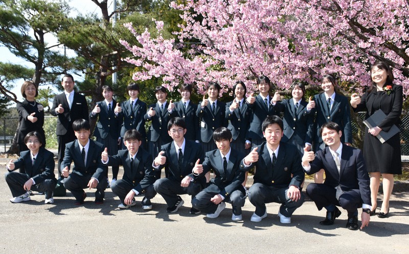 満開の桜の下で、親指で能登半島の形を作って記念撮影をする穴水高校の新入生と教職員ら＝いずれも石川県穴水町で、2026年4月8日午後2時16分、中尾卓英撮影