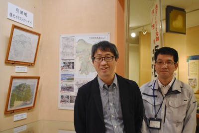 引田城を復元したイラスト2作品の展示コーナーのそばに立つ八木さん（左）と香川県東かがわ市教委の猪熊全徳さん＝同市歴史民俗資料館で2026年4月16日午後2時31分、佐々木雅彦撮影