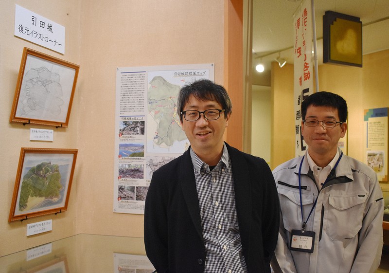 引田城を復元したイラスト2作品の展示コーナーのそばに立つ八木さん（左）と香川県東かがわ市教委の猪熊全徳さん＝同市歴史民俗資料館で2026年4月16日午後2時31分、佐々木雅彦撮影