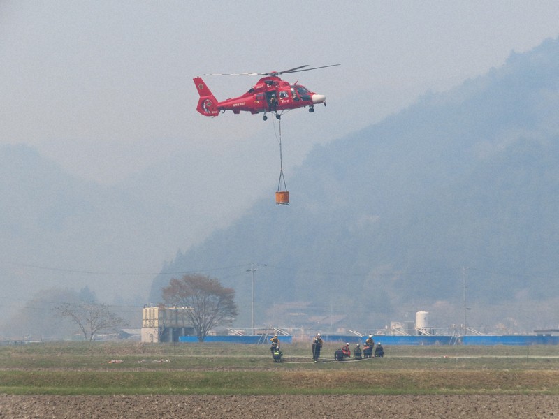 山林火災の煙が舞う中、川の水をつり下げて上昇するヘリコプター＝岩手県大槌町で2026年4月26日、工藤哲撮影