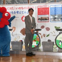 「いのちのはかり」に牛乳の模型を載せる山下真知事（右）とミャクミャク＝奈良市の平城宮跡歴史公園で、田辺泰裕撮影