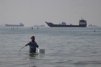 A man stands in the water, appearing to fish, as bulk carriers, cargo ships, and service vessels line the horizon in the Strait of Hormuz off Bandar Abbas, Iran, April 27, 2026.(Razieh Poudat/ISNA via AP)